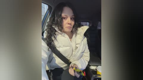 Video of a woman with long curly black hair, wearing a light-colored puffer jacket, seated in a car with a seatbelt across her chest. The background is dimly lit with a blurred view of the interior.