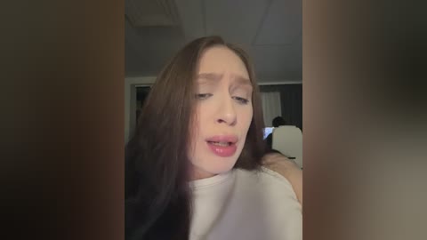 Video of a young woman with long brown hair, wearing a white top, looking pensive, indoors with blurred background.