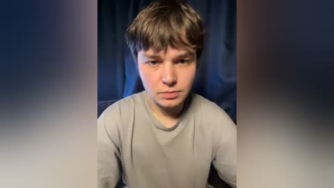 Video of a young boy with light brown hair, wearing a gray shirt, looking solemnly at the camera against a dark, blurred background.