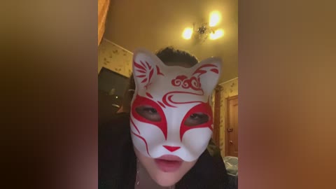 Video of a person wearing a white fox mask with red markings, standing in a dimly lit room with yellow wallpaper, wooden door, and chandelier.