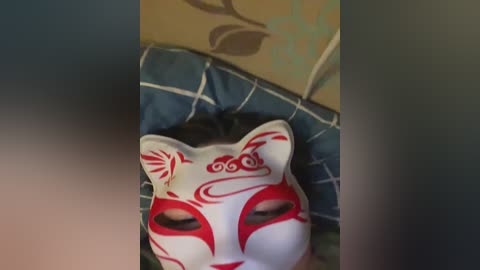 Video of a white mask with red and black tribal patterns, lying on a blue and green patterned quilt. The mask has a stylized fox face with large, expressive eyes.