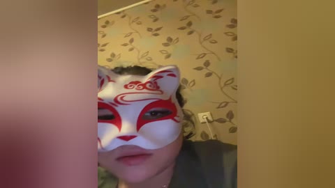 Video of a person wearing a white fox mask with red markings, in a room with beige wallpaper and floral pattern.