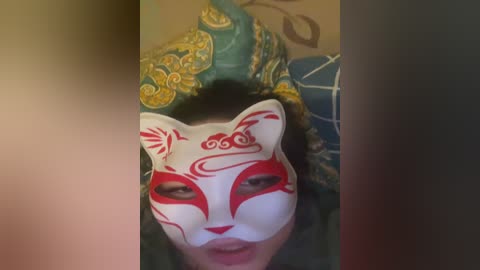 A video of a person with dark hair wearing a traditional white fox mask with red and gold patterns, positioned against a blurred background featuring blue and green fabric with intricate designs.