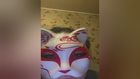 Video of a person wearing a white fox mask with red markings and intricate patterns, set against a beige wallpaper with floral designs. The person's face is partially visible, with dark hair, and the image is slightly blurred.