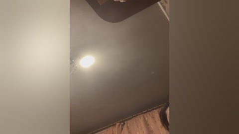 A video of a dimly lit ceiling with a bright, circular light bulb emitting a soft glow. The ceiling is a dull beige, and the light casts a warm, soft light.