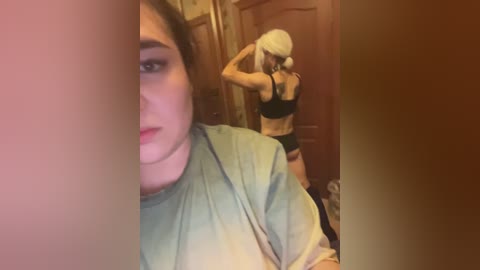 Video of a woman with light skin and long blonde hair, wearing a gray shirt, standing in a dimly lit room with a wooden door and brown walls. Another woman, similarly dressed, is seen in the background.