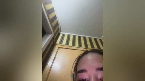 Video of a woman with light brown skin and long, wet hair, partially visible from the neck up, looking down. The background features a corner of a room with yellow-striped wallpaper.