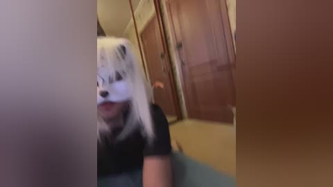 A blurry video shows a person in a black t-shirt and a white mask resembling a panda, standing in a hallway with wooden doors and beige walls.