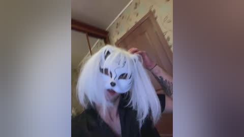 A video shows a person with long, white hair wearing a white mask resembling a dog's face, holding their hair, in a room with light-colored walls and a wooden door.