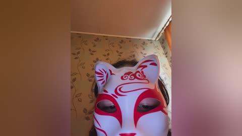 Video of a person wearing a white and red fox mask, standing in a room with beige wallpaper adorned with floral patterns. The mask has intricate red markings and a serene expression.