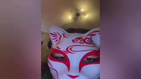 A video of a person wearing a white fox mask with red tribal patterns, set against a dimly lit room with a ceiling fan visible.