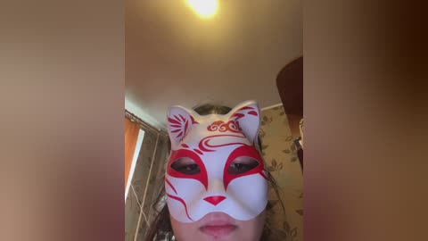 A video captures a person wearing a white, red-patterned fox mask indoors, with a soft, warm light illuminating the ceiling. The background includes a patterned wall and a glimpse of a wooden chair.