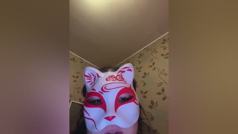 A video of a person wearing a white fox mask with red markings, standing in a room with beige walls and floral wallpaper. The image is taken from a low angle, emphasizing the mask's intricate design.