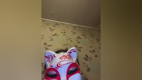 Video of a white mask with red and black patterns, worn by a person, standing in a beige room with floral wallpaper.