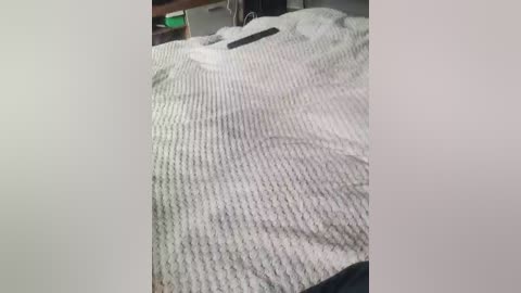 A video of a white, textured blanket with a dark-colored pillow on a bed. A black smartphone is placed on the blanket, slightly out of focus. The background includes a blurred green object and a wooden surface. The image has a soft, natural lighting.