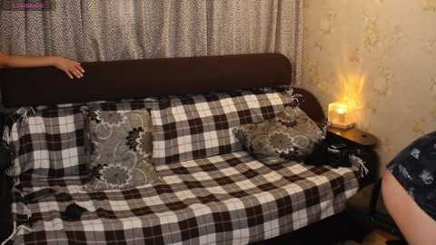 Video of a cozy, vintage-style living room with a plaid-covered couch, floral-patterned pillows, and a warm-toned lamp on a side table, set against a patterned wallpaper background.