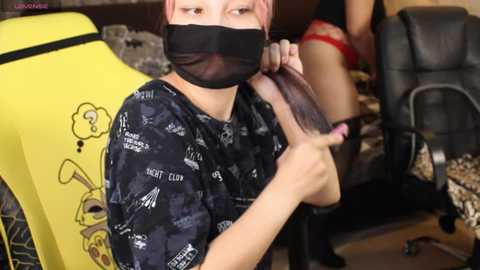 Video of a person with light skin and short pink hair, wearing a black mask and a dark shirt with text, trimming hair in a salon, with a yellow chair and another person in red underwear visible in the background.