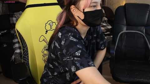 Video of a young woman with pink hair, wearing a black face mask, sitting in a yellow gaming chair with a cartoon rabbit, in a dimly lit room.