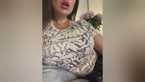 A video of a light-skinned woman with long brown hair, wearing a tight, patterned top that emphasizes her large breasts. She's indoors, with a vase of purple flowers in the background.