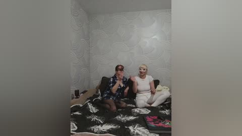 Video of two women on a bed in a small, dimly lit room with white, patterned wallpaper. One woman wears a blue plaid shirt, the other a light pink top.