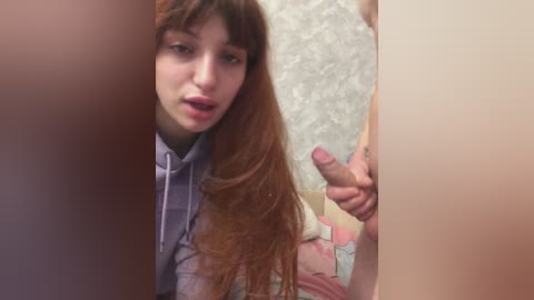 A video of a young woman with long, wavy red hair, wearing a light blue hoodie, standing in a bathroom with floral wallpaper, holding an erect penis in front of her.