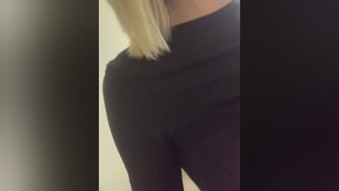 Video of a woman's torso wearing a black high-waisted turtleneck and black leggings. Her straight, blonde hair partially obscures her face. The background is a blurred gradient from light to dark.
