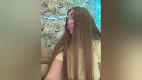 A video of a young woman with long, straight brown hair, wearing a yellow t-shirt, standing indoors against a floral-patterned wallpaper. She appears relaxed, with a slight smile.