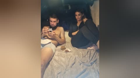 Video of a young, shirtless man with a baby, and a woman sitting on a bed, both engaged with smartphones.