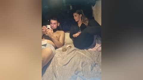 Video of a man and woman in bed, man topless with white shorts, woman in black hoodie and jeans, both looking at phone, gray blanket.