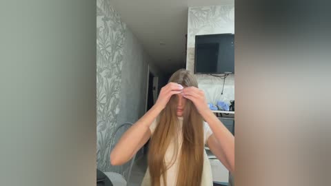 Video of a young woman with long, straight brown hair, holding her hair up, standing in a modern, minimalist bedroom with light gray wallpaper and a flat-screen TV on the wall.