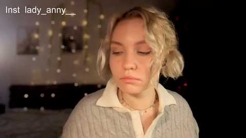 Video of a young woman with shoulder-length blonde hair, wearing a light gray sweater, standing in a dimly lit room with string lights and a bed in the background.