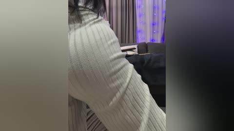 Video of a woman's lower body in a light gray ribbed knit dress, accentuating her curvy figure, standing indoors with a sofa and curtains in the background.