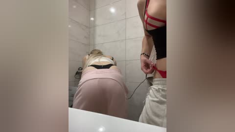 Video of a bathroom scene: a blonde woman in a black bra and pink shorts bent over a toilet, a woman in a red bra and white shorts standing behind her.
