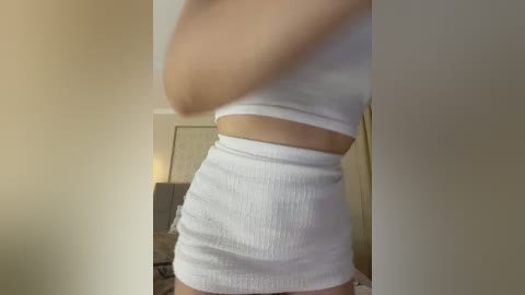 Video of a light-skinned woman in a white ribbed crop top and matching high-waisted shorts, captured from the side in a dimly lit room with beige walls.