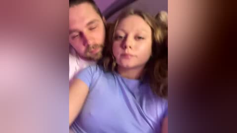 A video captures a young, fair-skinned woman with light brown hair and a man with short dark hair and a beard, both in casual attire, appearing relaxed in a dimly lit room.