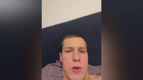 A video of a young man with light skin and short brown hair, lying on a bed with a purple and white patterned sheet. He appears to be in a state of discomfort or pain, with a slightly open mouth and furrowed brow.
