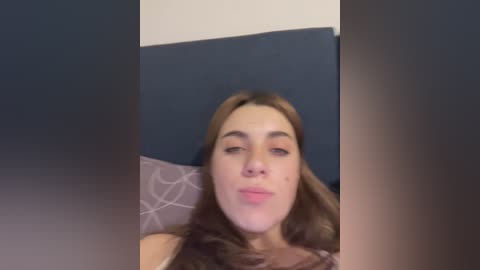 Video of a young woman with long, light brown hair, light skin, and freckles, wearing a brown top, lying on a bed with dark blue and white patterned pillows.