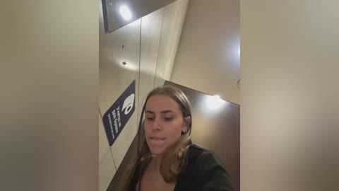 Video of a blonde woman with long hair, wearing a black jacket, standing in a dimly lit, beige-tiled hallway with a reflective ceiling and a circular light fixture.