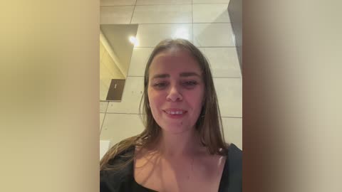 A video of a young woman with long brown hair, wearing a black top, smiling, standing in a tiled bathroom with beige walls and a mirror.