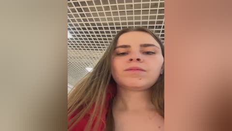Video of a young Caucasian woman with straight, shoulder-length blonde hair, light skin, and neutral expression, taken in an indoor setting with white grid ceiling tiles and beige walls.