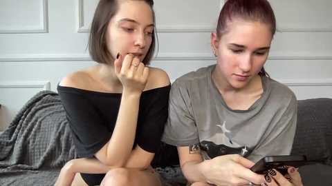 Video of two young women, one with short brown hair, black off-shoulder top, and the other with red hair, gray tee, both sitting on a gray couch, engrossed in smartphones.