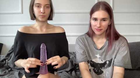 Video of two young women with light skin and straight hair, one with short bob and black off-shoulder top, the other with long red hair and gray t-shirt. They hold a purple dildo. Background features a white paneled wall and a gray couch.
