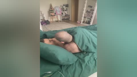 Video of a naked man with a shaved head and light skin, lying on a green bedspread in a messy bedroom with cluttered shelves and a cat.