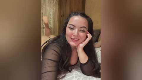 Video of a smiling woman with long black hair, wearing a sheer black top, lying on a bed, legs crossed, in a cozy, warmly lit bedroom.