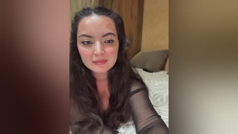 A video of a light-skinned woman with long, dark hair and a makeup look, smiling, in a bed with a white patterned blanket, beige walls, and a brown door frame.