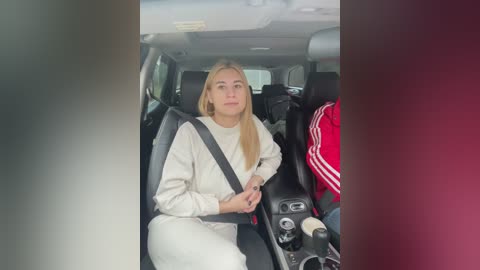 Video of a young blonde woman wearing a white sweater and pants, seated in a car with a red jacket and black seatbelt visible.