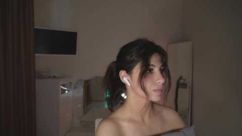 A video of a woman with dark hair, wearing white earbuds, sitting in a dimly-lit, modern room.