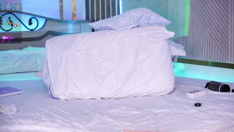 A video of a neatly made bed with white linens and a plush pillow, a remote control, and a laptop on the bed. The background shows a modern, dimly lit room with a sleek, black metal headboard.