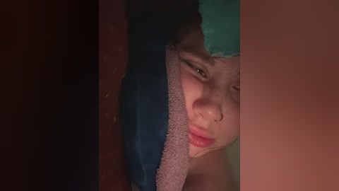 A video of a young woman with light brown skin and curly hair, partially covered by a blue towel, seen through a narrow slit. Her face is partially visible, displaying a pensive expression.