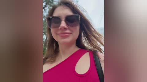Video of a Caucasian woman with light skin, long brown hair, and large, dark sunglasses, wearing a bright pink one-shoulder top. She smiles slightly. Background is blurred, suggesting an outdoor setting.